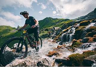 Vététiste roulant sur un sentier rocailleux le long d’un ruisseau de montagne dans un paysage alpin près d’Arosa, entouré de montagnes verdoyantes et d’un ciel bleu.